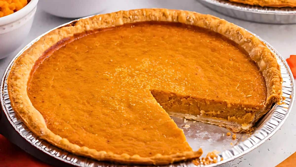 A golden-brown pumpkin pie with a slice missing, revealing a smooth, creamy interior. The pie sits in a metal foil pan on a light-colored surface.