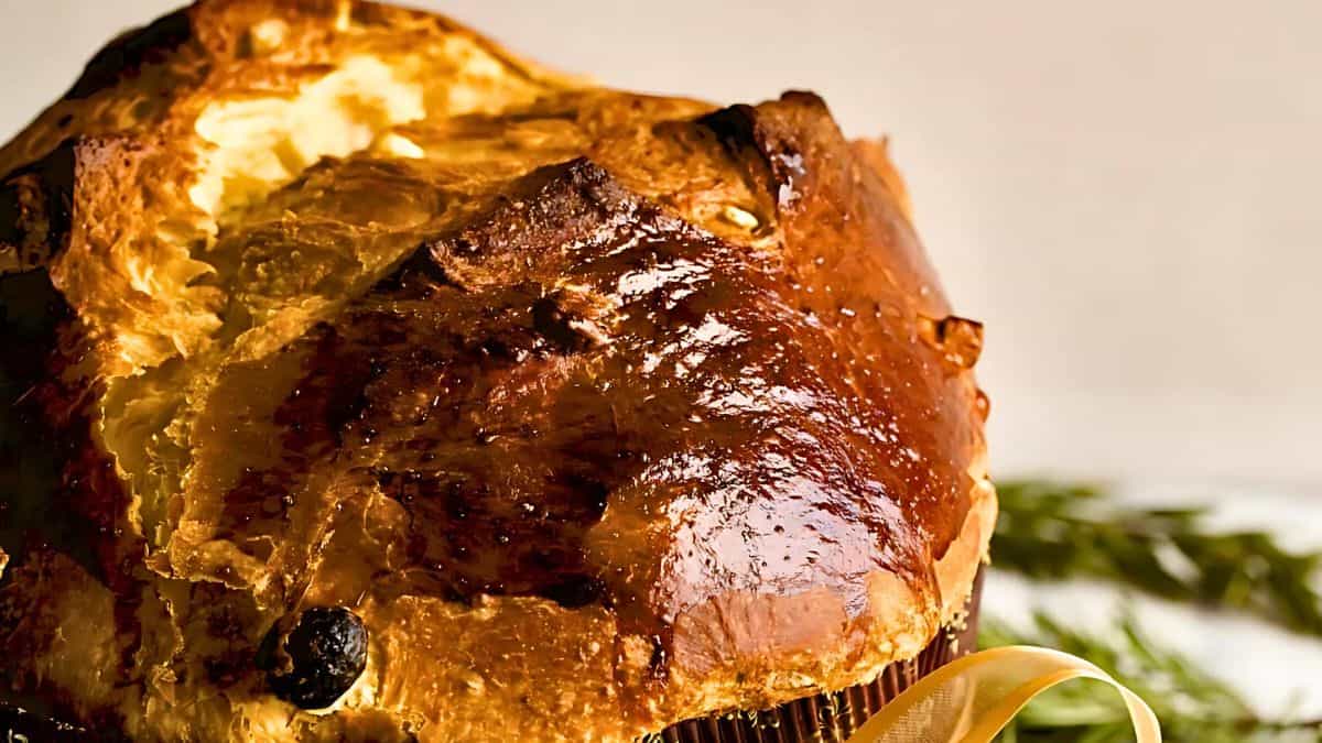 Close-up of a golden-brown panettone with a glossy crust, set against a neutral background. A sprig of greenery with a ribbon peeks out from the side.