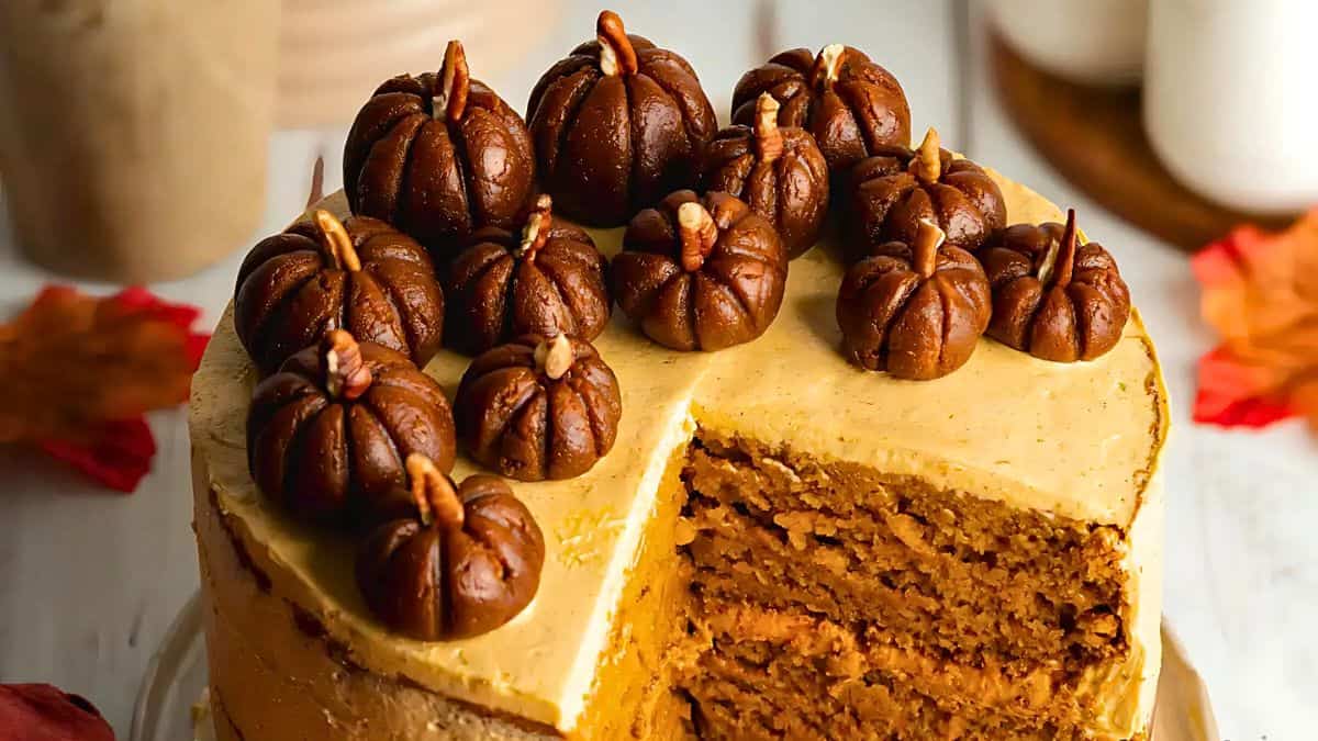 A pumpkin spice cake with a slice removed, showcasing layered texture. The cake is topped with chocolate icing shaped like small pumpkins. In the background are blurred autumn leaves and mugs, creating a cozy autumn atmosphere.