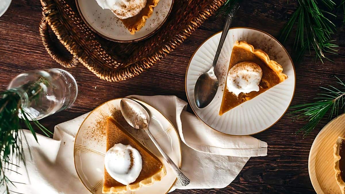 Top view of two slices of pumpkin pie topped with whipped cream on plates, accompanied by spoons. The setting includes a rustic wooden table and a woven basket, with green pine branches adding a festive touch.