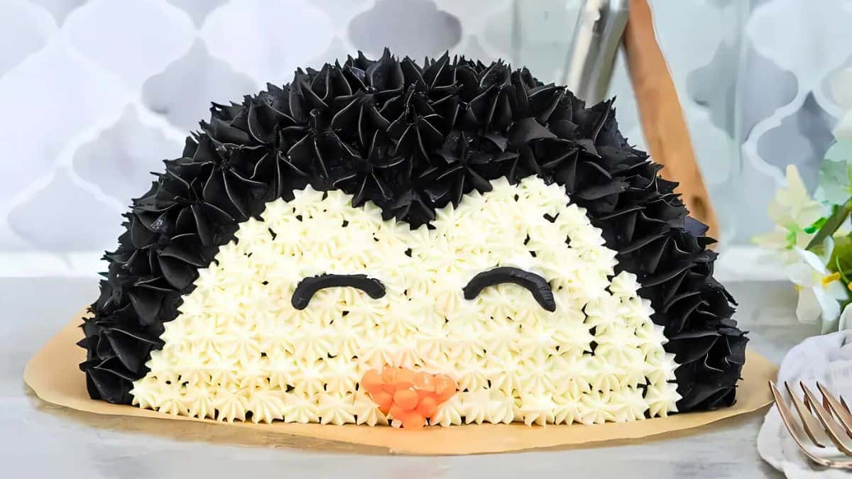 A cake shaped like a cute penguin face is adorned with black and white frosting. The penguin has closed eyes and an orange beak, elegantly decorated to appear realistic. The background shows a blurred, patterned surface.