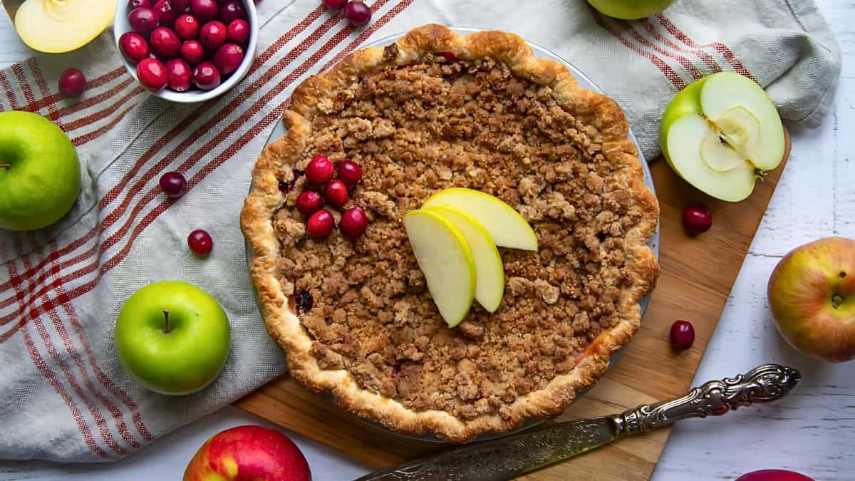 A crumble pie topped with apple slices and cranberries sits on a wooden board next to green apples, cranberries, and a striped cloth.