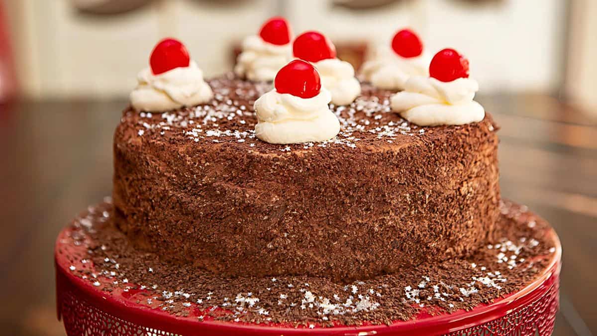 A decadent chocolate cake topped with whipped cream and cherries, set on a decorative red cake stand. The cake's surface is sprinkled with powdered sugar and chocolate shavings.