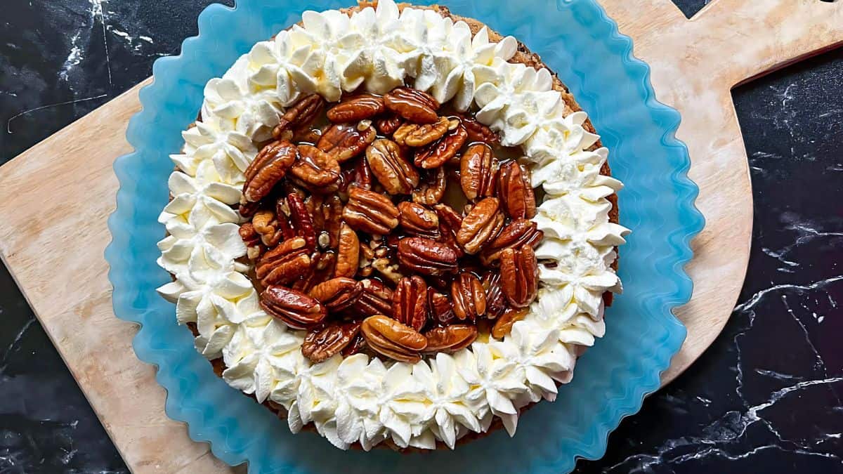 A round pecan pie topped with whipped cream swirls and glazed pecans sits on a blue plate with a wavy edge, placed on a wooden board against a dark marble background.