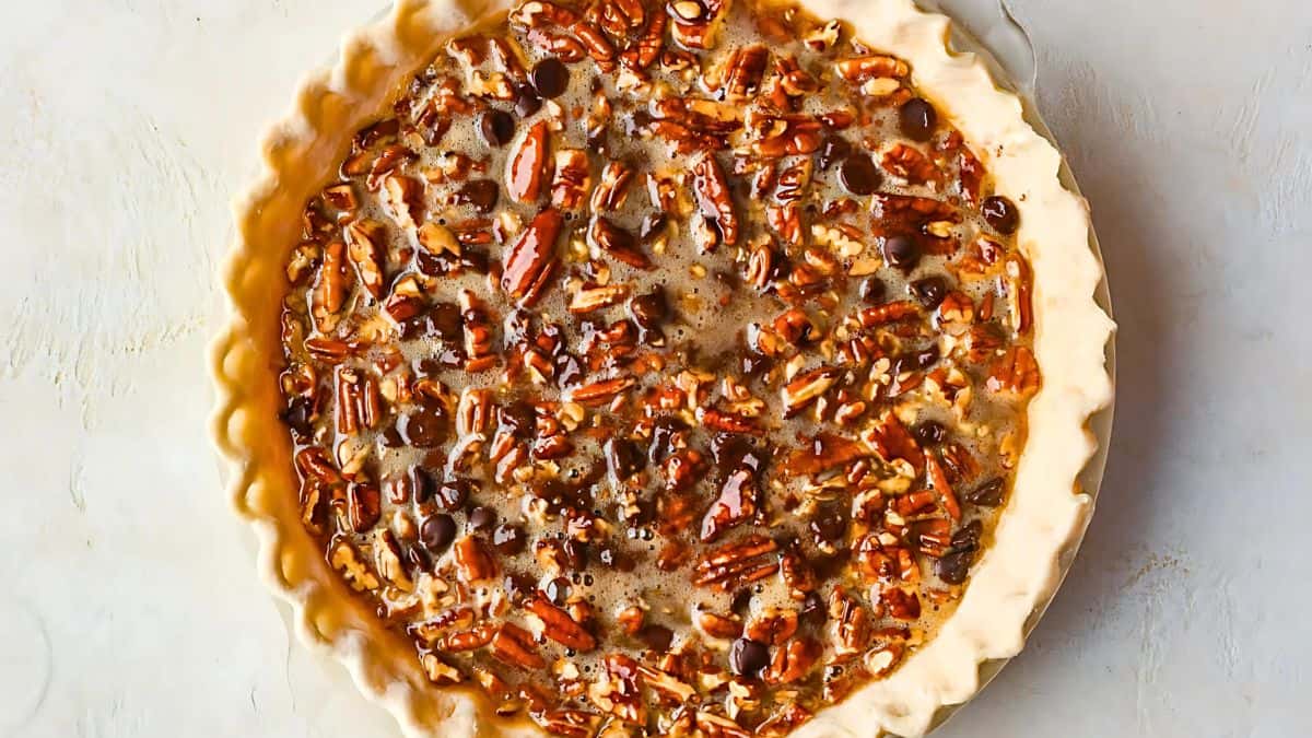 A pie with a golden crust is filled with pecans and chocolate chips, creating a mouthwatering dessert. The surface of the pie is glossy and textured, reflecting light, and it's set against a light-colored background.