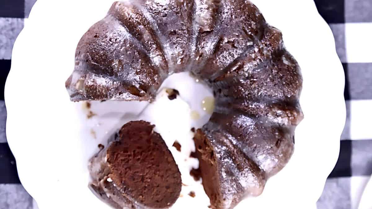 A chocolate bundt cake with a glossy glaze on top, placed on a white plate. One slice is cut and positioned at the center of the cake, revealing its rich, moist interior. The plate rests on a checkered black and white tablecloth.