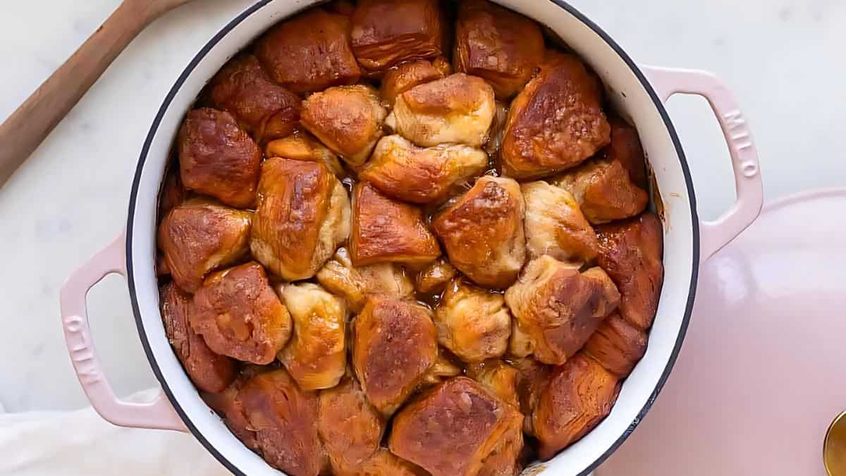 A pink pot filled with baked monkey bread, featuring golden-brown, fluffy dough pieces. The pot rests on a light surface next to a wooden spoon.