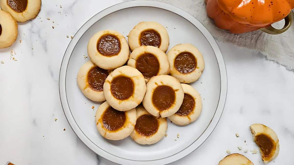 A plate of thumbprint cookies filled with caramel sits on a white marble surface. Crumbs are scattered around, and a ceramic orange mug with a gold handle is partially visible in the top right corner.