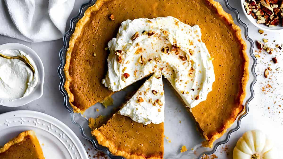 A top view of a pumpkin pie with creamy white topping and sprinkled nuts. One slice is cut and partially removed. The pie sits on a clear glass dish, surrounded by whipped cream and chopped nuts in small bowls on a marbled surface.