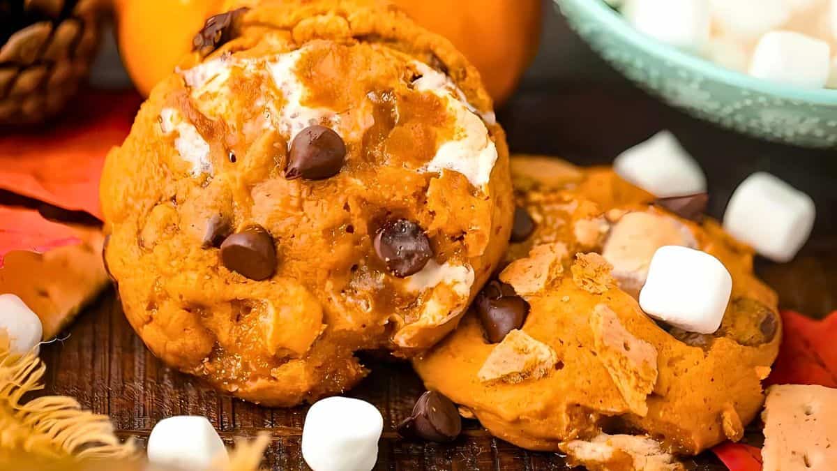 Two pumpkin chocolate chip cookies topped with marshmallows sit on a wooden surface, surrounded by small marshmallows and autumn leaves. A bowl with more marshmallows is visible in the background.