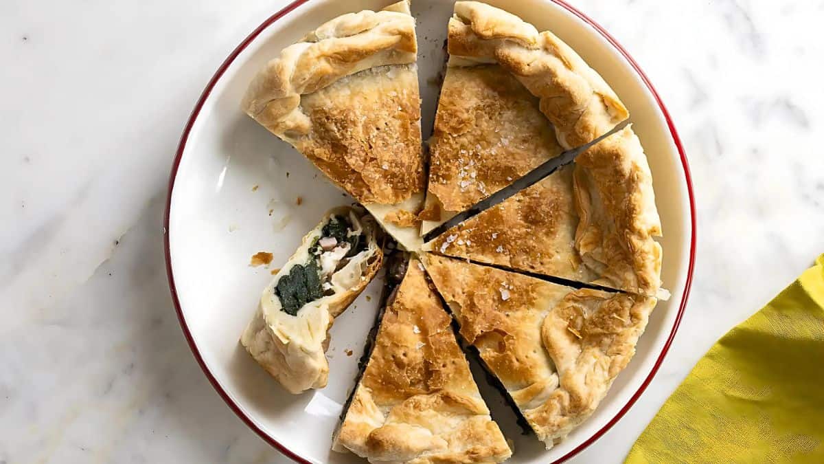 A sliced spinach and cheese pie is served on a white plate with a red rim. One piece is turned to reveal the filling of dark green spinach and white cheese. The crust is golden brown and flaky. A yellow napkin is partially visible on the right.