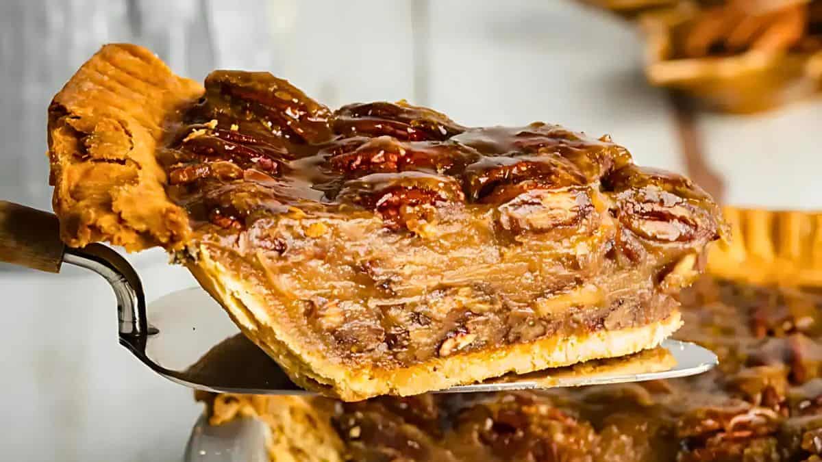 A slice of pecan pie being lifted from the pie dish. The slice showcases a flaky crust, gooey caramel filling, and a topping of whole pecans, all in rich, warm tones.