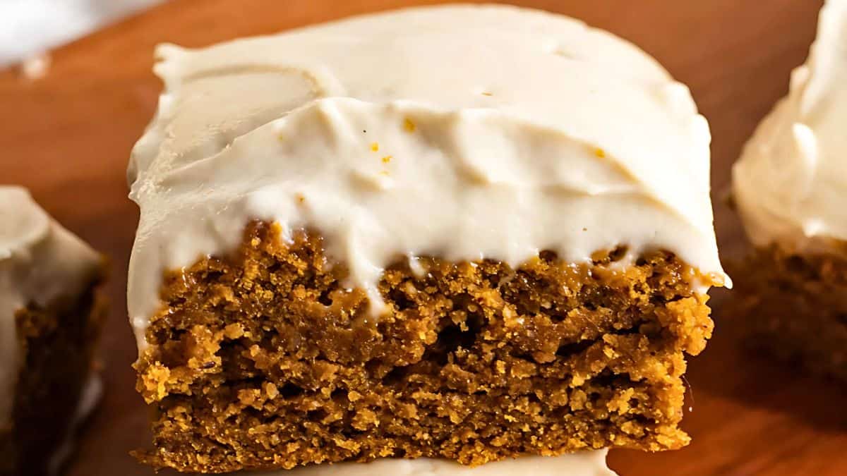A close-up of a slice of moist, spiced cake topped with creamy frosting. The cake has a rich, golden-brown color and a light, fluffy texture, with the frosting generously spread on top. It rests on a wooden surface.