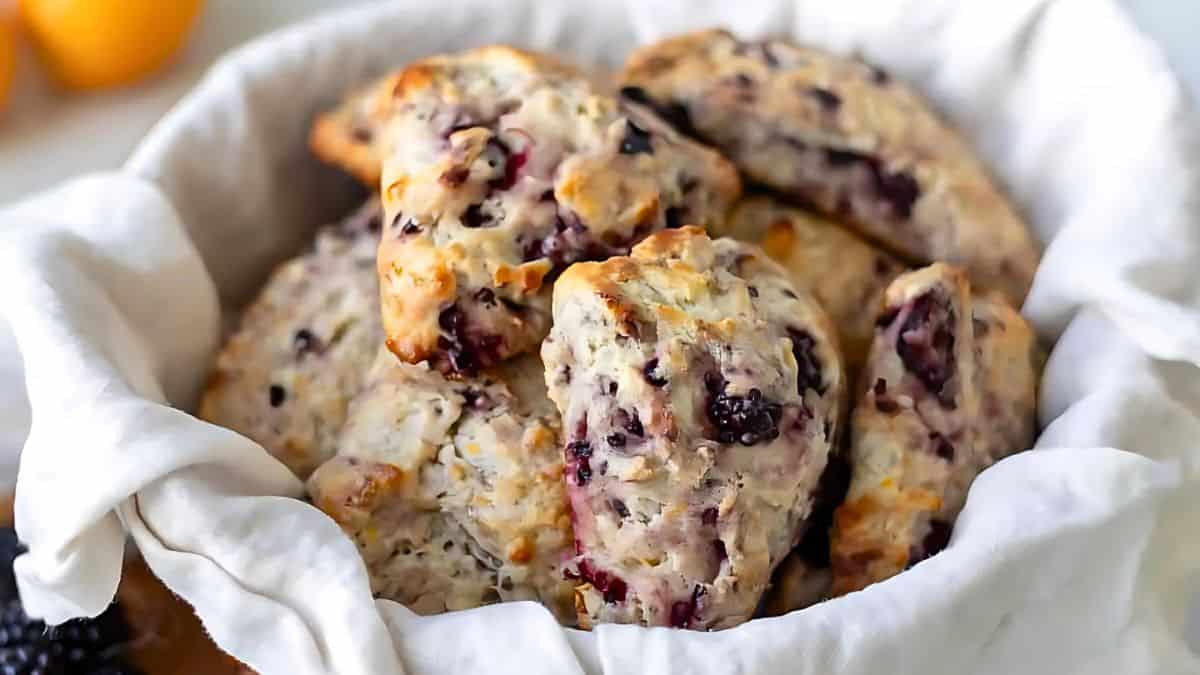 A basket lined with a white cloth is filled with freshly baked scones. The scones have a golden-brown exterior with visible berries and flaky textures. In the background, there are hints of citrus fruits.