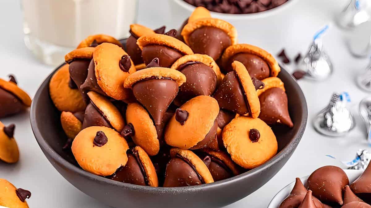 A bowl filled with chocolate and peanut butter acorn-shaped treats made from chocolate kisses, mini peanut butter cookies, and chocolate chips. A glass of milk and a bowl of chocolate chips are in the background.