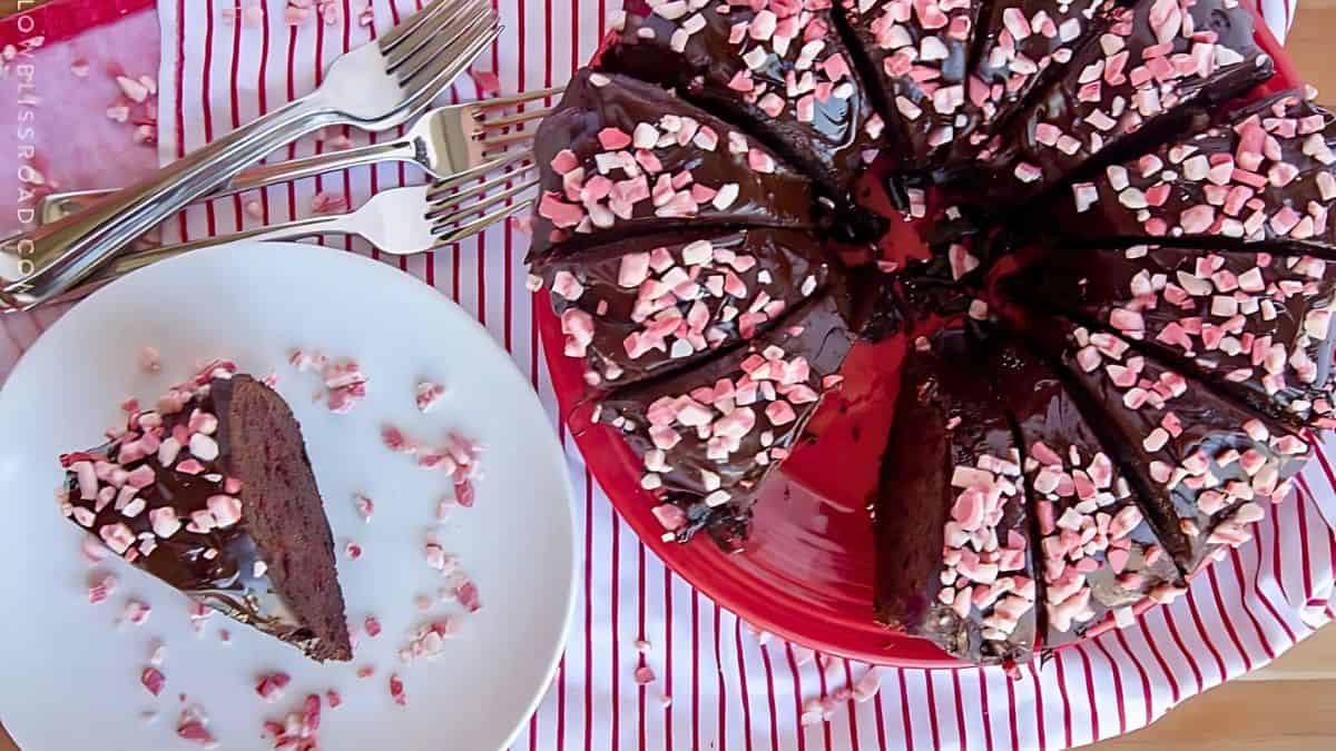 A chocolate cake with a rich glaze is topped with pink and white heart-shaped sprinkles. A slice is served on a white plate with a scattering of sprinkles. Three forks rest beside the plate on a red and white striped cloth.