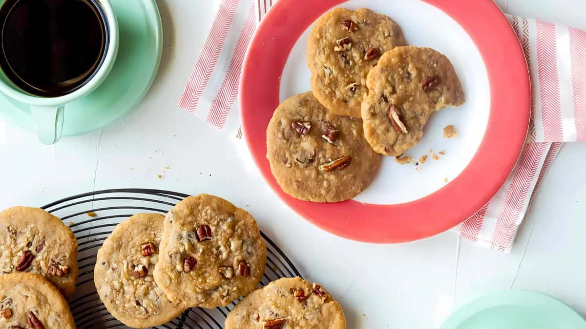 A table with plates of pecan cookies, one on a red and white plate, and another on a cooling rack. A cup of black coffee sits on a green saucer, next to red and white striped napkins.