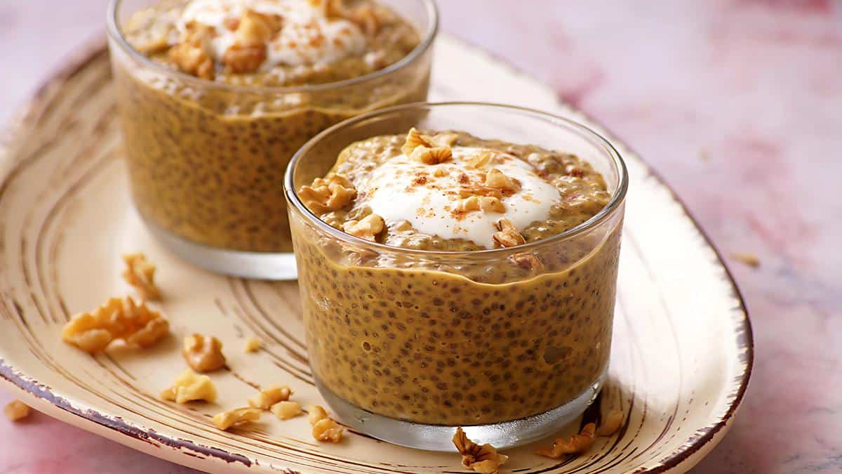 Two glass cups filled with chia seed pudding, topped with yogurt and sprinkled with chopped walnuts, are placed on a rustic platter. The background is softly blurred pink.