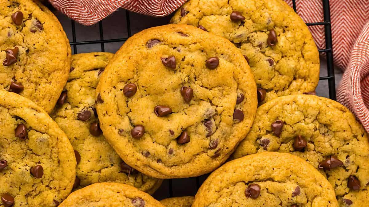 A close-up of freshly baked chocolate chip cookies on a cooling rack, surrounded by a red and white striped cloth. The cookies are golden brown and studded with chocolate chips.