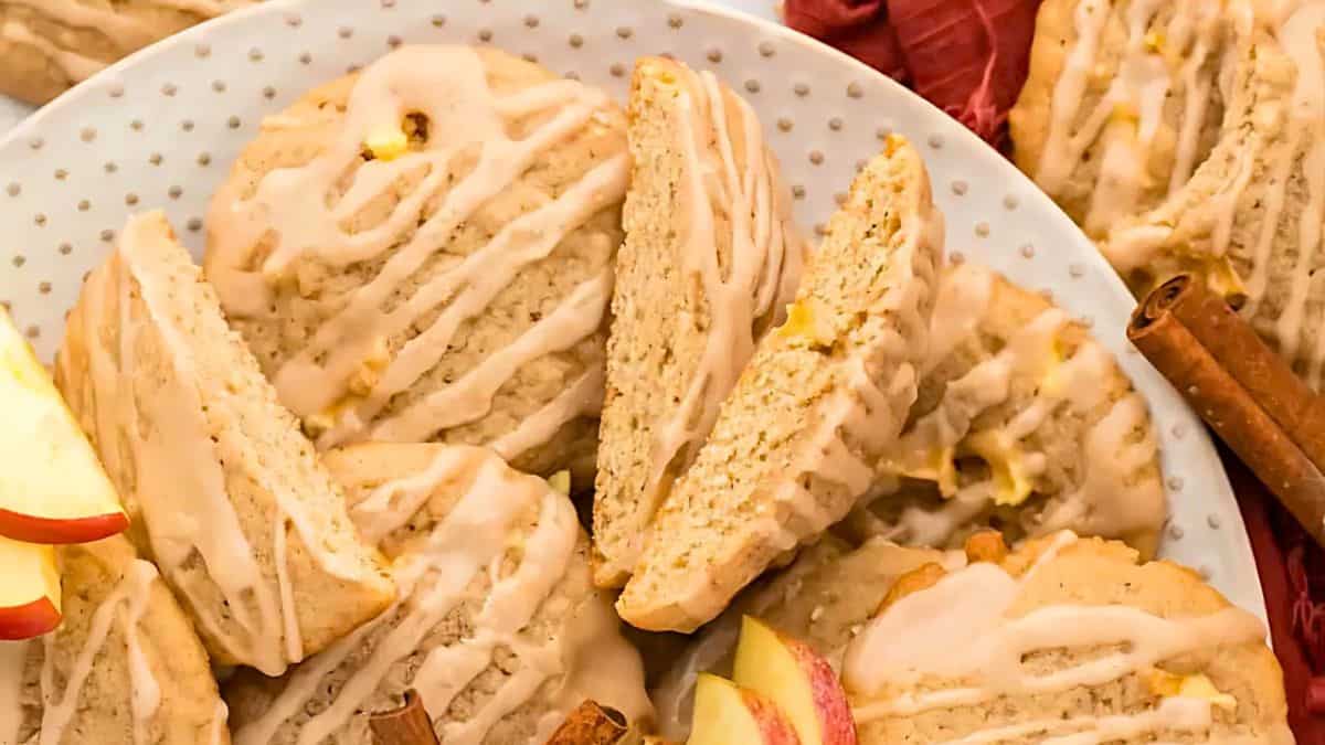 A plate of iced apple cinnamon scones with a few apple slices and cinnamon sticks. The scones are drizzled with icing and stacked on a white polka dot plate. A red cloth is seen in the background.
