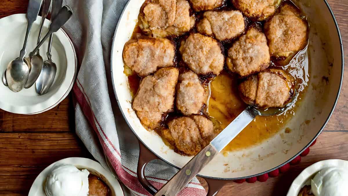 A top view of a large baked dish filled with golden brown biscuit-like toppings arranged over a caramelized sauce. A serving spatula is resting inside. Two bowls with scoops of vanilla ice cream and three spoons are placed nearby on a wooden table.