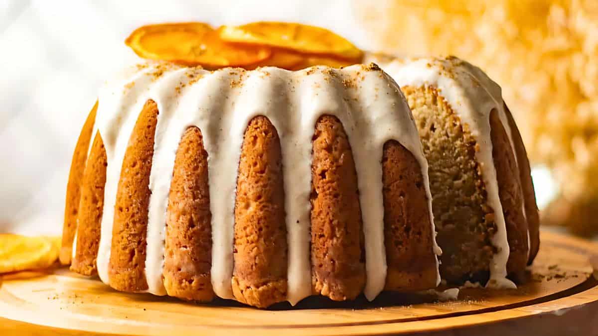 A bundt cake with white icing drips over the top, topped with dried orange slices. The cake is placed on a wooden board, warmly lit, with a blurred background featuring soft, golden light and floral elements.