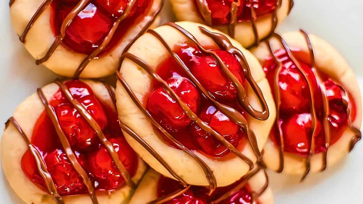 Close-up of cookies topped with red cherries and drizzled with chocolate. The cookies have a golden-brown crust, and the glossy cherries are vibrant and juicy, complemented by the dark chocolate drizzle.
