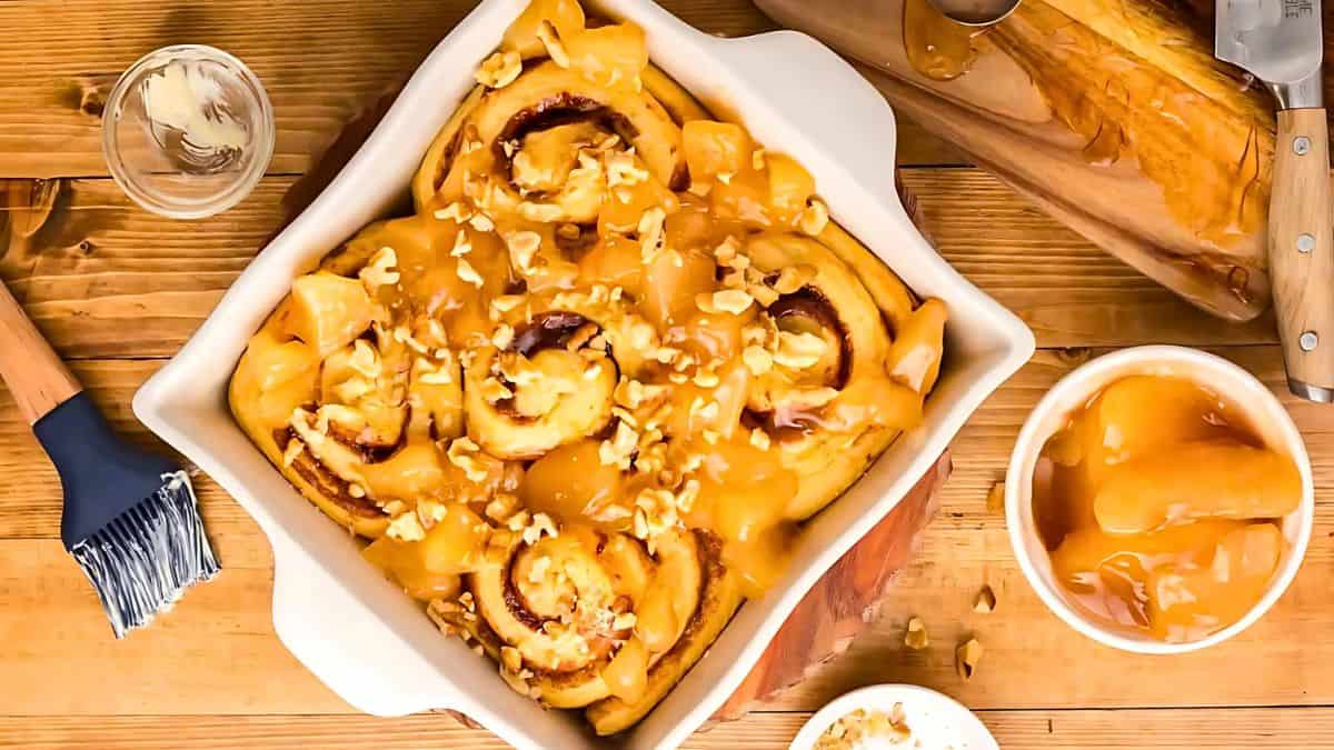 Freshly baked cinnamon rolls in a square white dish are drizzled with caramel sauce and topped with chopped nuts. A small bowl with caramel topping and cooking utensils are nearby on a wooden surface.