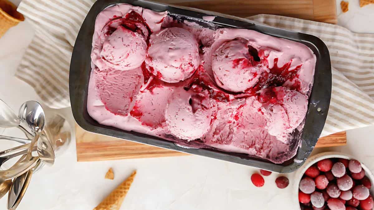 A baking tray filled with scoops of raspberry ice cream, garnished with raspberry sauce, sits on a wooden board. On the side, there are metal spoons, a striped cloth, bits of waffle cone, and a bowl of frozen raspberries.