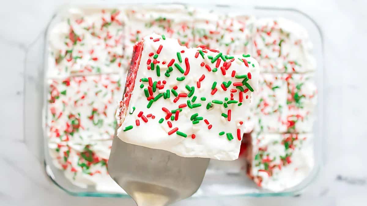 A piece of red velvet cake with white frosting and red and green sprinkles is lifted on a spatula. The cake below sits in a glass baking dish, also topped with frosting and sprinkles.