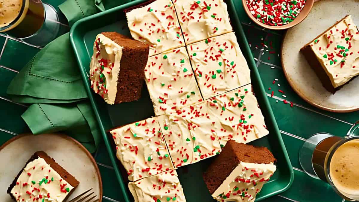 A tray of frosted chocolate cake squares is topped with red and green sprinkles. Some slices are served on plates nearby. The scene is set on a green tablecloth with a green napkin and cups of coffee.