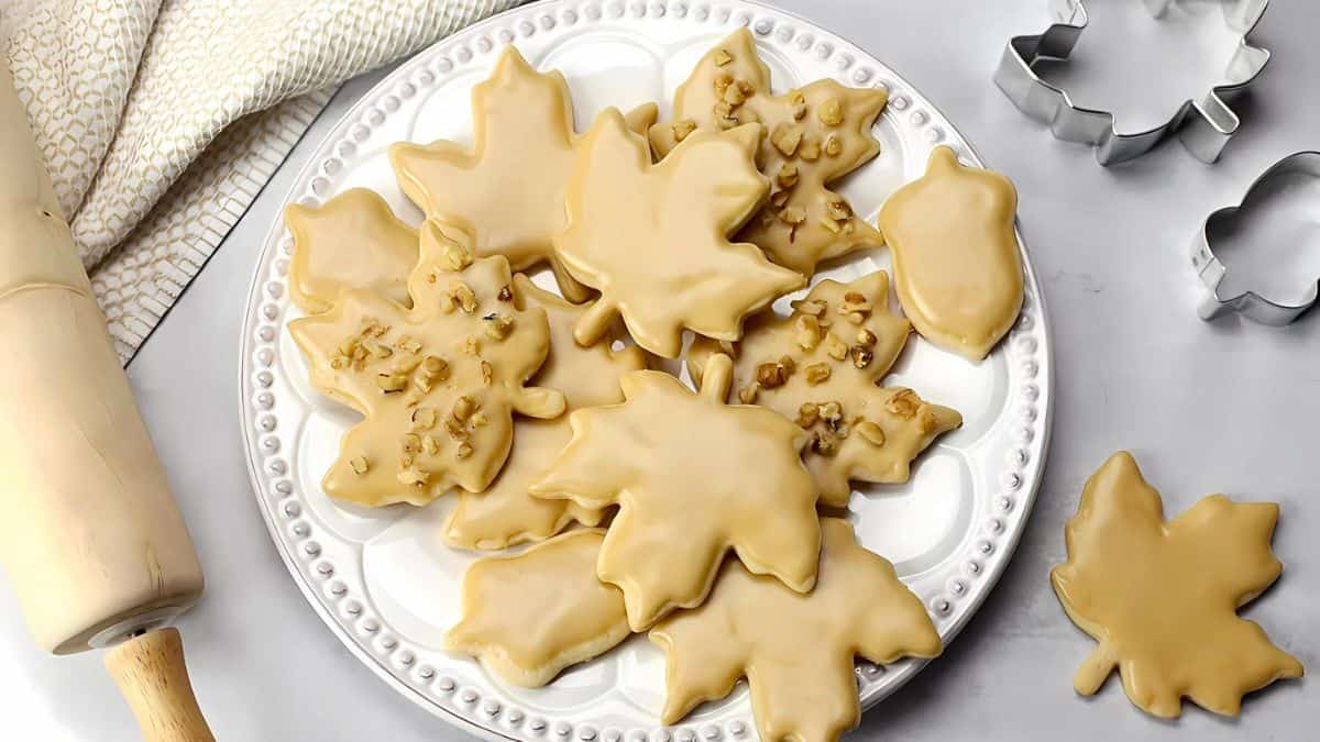 A plate of maple leaf-shaped cookies decorated with beige icing and small nut pieces. Nearby are autumn-themed cookie cutters, a rolling pin, and a textured kitchen towel on a white surface.