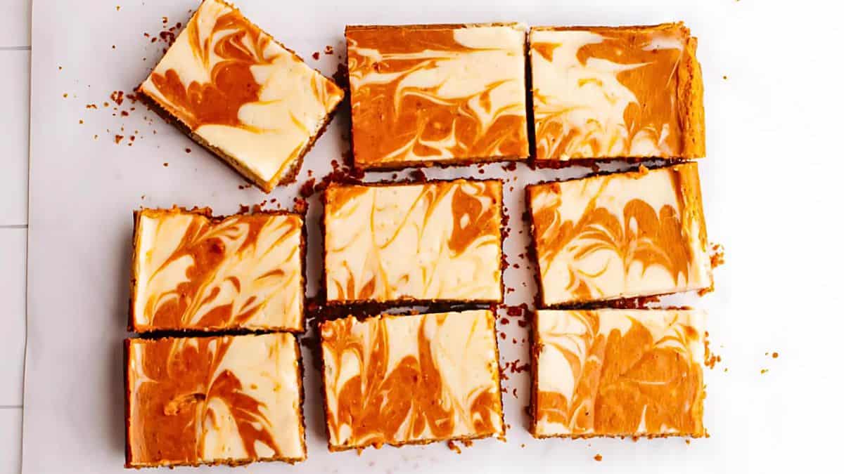 Top view of eight pumpkin swirl cheesecake bars arranged on a white surface. The bars have a marbled design with orange and cream colors, each cut into rectangular pieces. Crumbs are scattered around them.