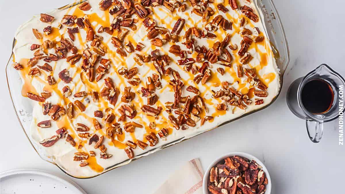 A rectangular dessert with creamy white frosting topped with pecan pieces and caramel drizzle in a glass dish. Nearby are a bowl of pecans and a small pitcher of syrup on a white surface.