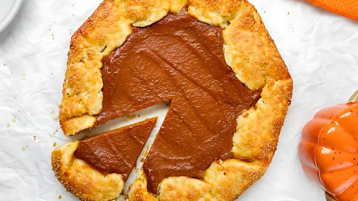 A rustic pumpkin galette with a slice cut out, showing its rich, smooth filling. The golden-brown crust is flaky and sprinkled with sugar, providing a textured edge. It rests on white parchment paper. An orange item is partially visible to the side.