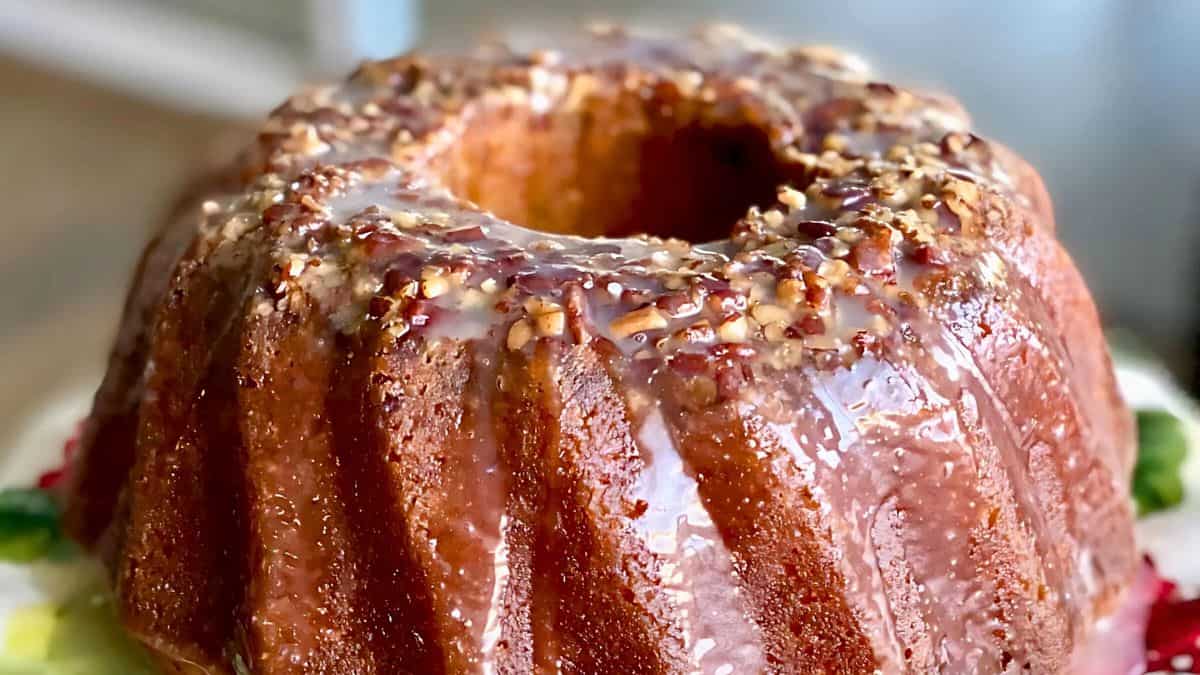 A close-up of a Bundt cake with a glossy glaze drizzled over the top, garnished with chopped nuts. The cake has a golden-brown color and is set on a floral-patterned plate.