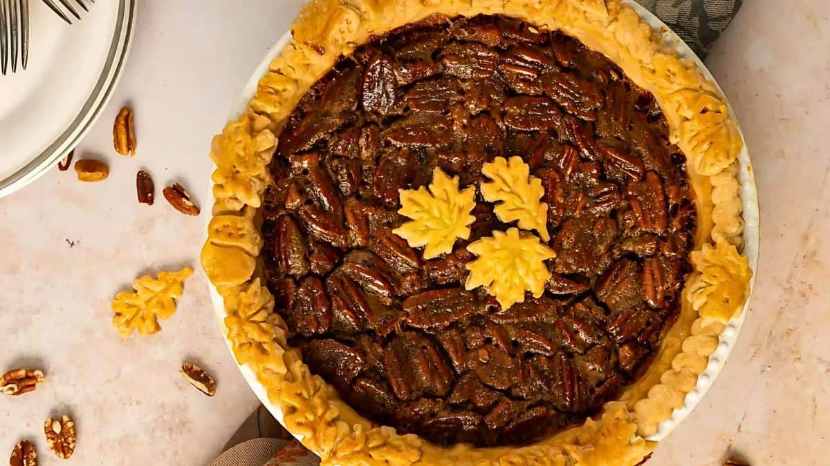 A whole pecan pie with a decorative crust featuring leaf-shaped pastry cutouts. Several pecans and leaf decorations are scattered around the pie on a light-colored surface. A stack of plates and forks is partially visible in the top left corner.