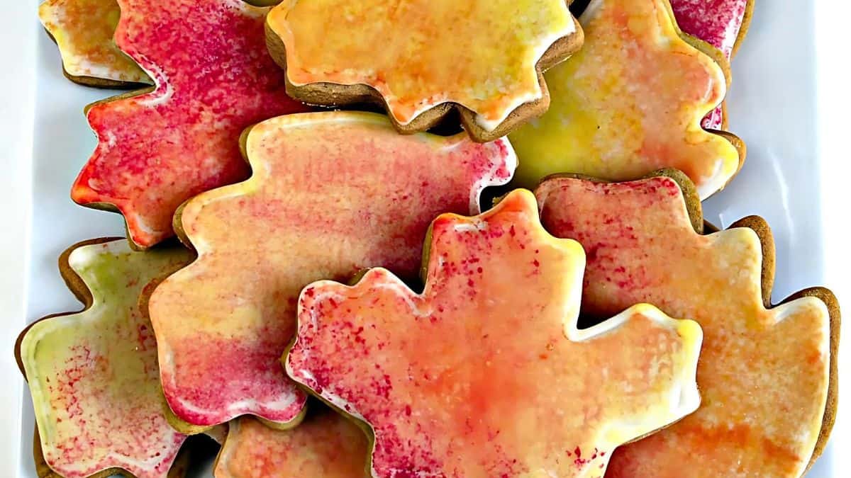 Plate of maple leaf-shaped cookies glazed in autumn colors of red, orange, and yellow. The cookies overlap each other, showcasing a vibrant seasonal theme.