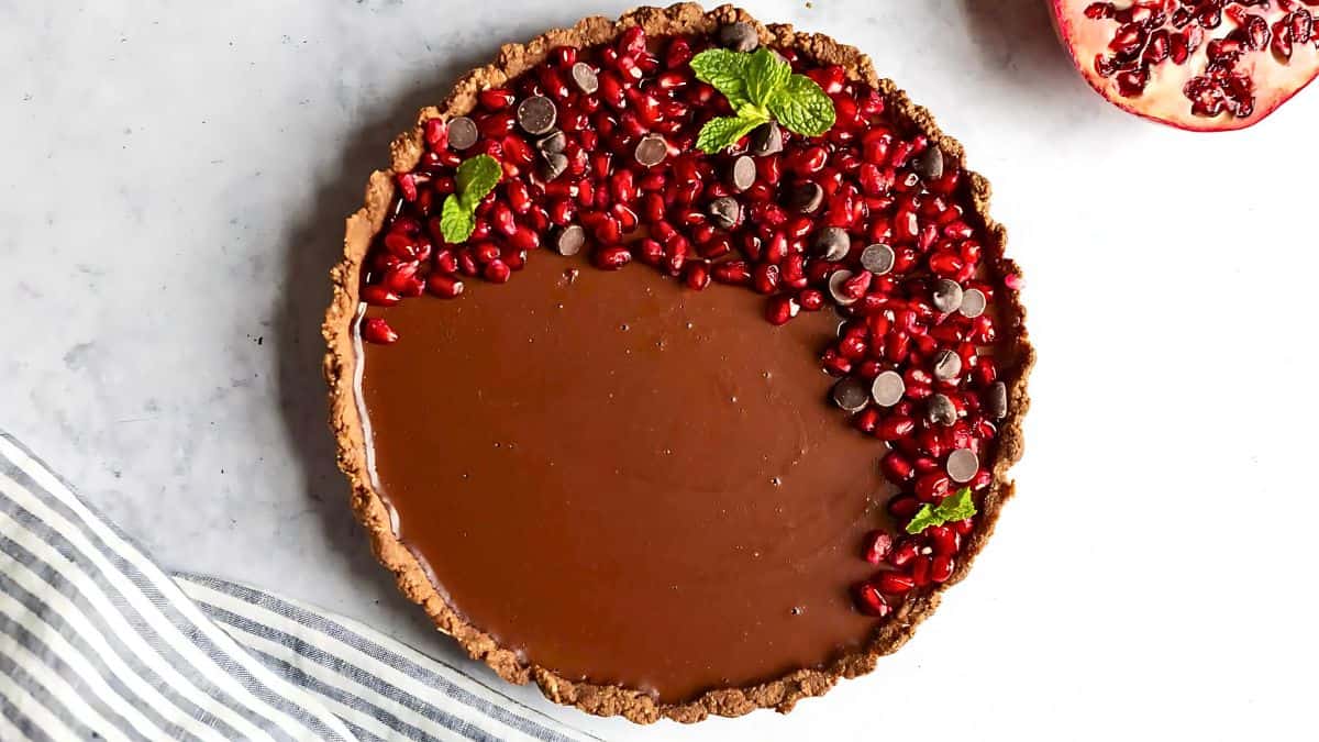 A chocolate tart topped with pomegranate seeds, chocolate chips, and mint leaves on a light marble surface. A striped cloth is partially visible at the bottom left, and a cut pomegranate is on the top right.