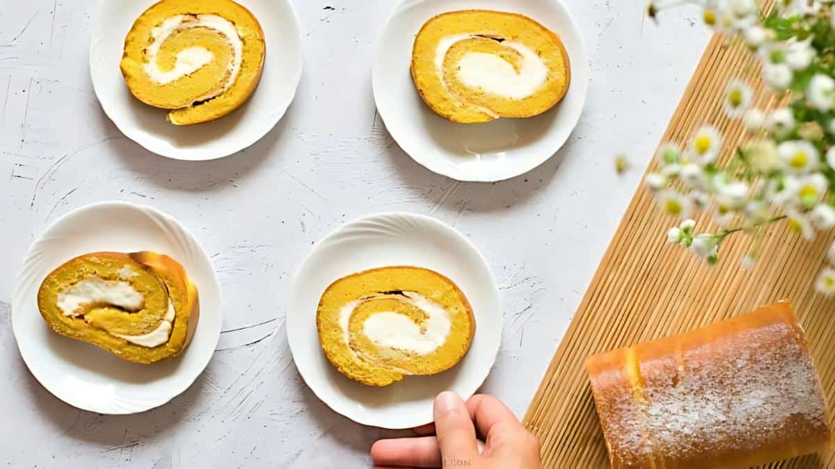 Three slices of a Swiss roll cake with a cream swirl on small white plates are arranged on a white surface. A whole cake is nearby on a wooden board. A hand reaches for one plate, and flowers are in the top right corner.
