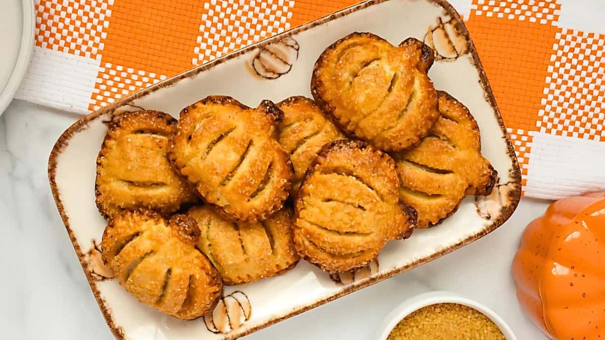 A plate of oval-shaped cookies with a golden-brown crust, displayed on a beige rectangular dish. The cookies have slits on top. The dish is on a surface with an orange and white checkered cloth underneath.