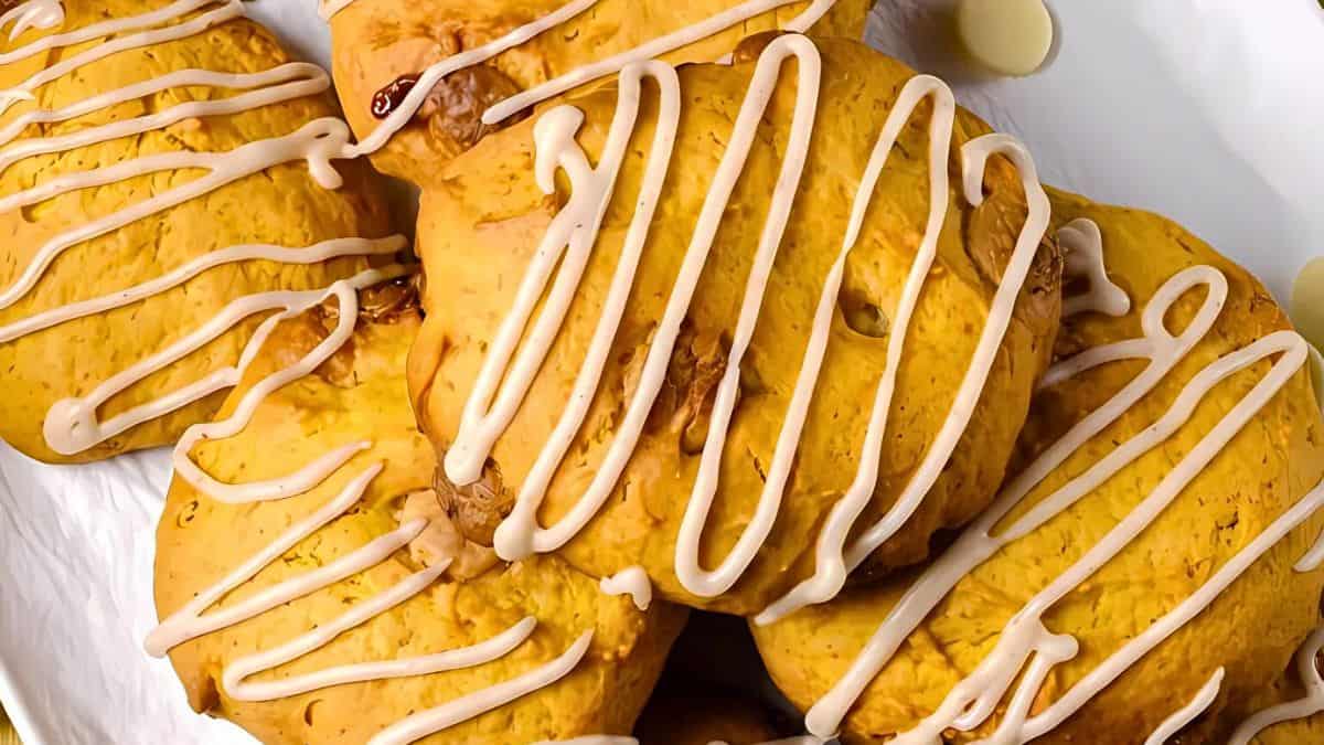 A close-up of pumpkin cookies drizzled with icing, stacked on a white surface. The cookies are round and have a light golden-brown color with visible nuts. The icing forms decorative lines across the tops of the cookies.