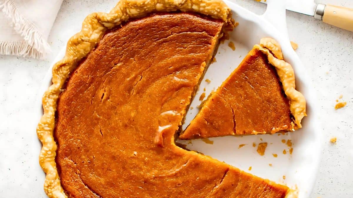 A whole sweet potato pie with a golden-brown crust in a white pie dish. One slice is cut and slightly removed, revealing a smooth and creamy orange filling. A pie server is visible nearby.