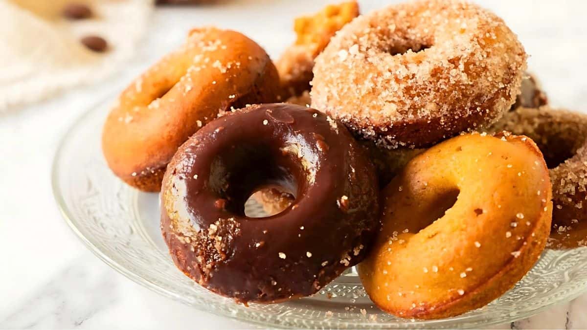 A plate of assorted donuts featuring sugar-coated and chocolate-glazed varieties, stacked together on a glass plate. The donuts have a golden brown hue, creating an appetizing display. The background is softly blurred.