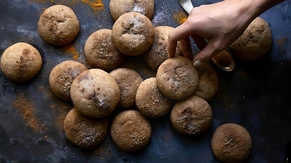 A hand reaches to pick up a snickerdoodle cookie from a scattered pile on a dark surface. The cookies are dusted with cinnamon sugar, creating a warm and inviting scene.