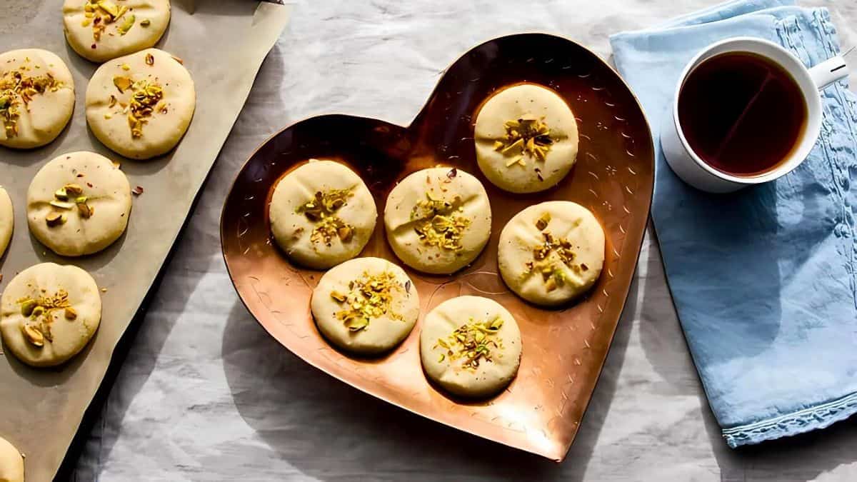 Heart-shaped tray with round cookies topped with pistachios, accompanied by a cup of tea on a blue napkin. More cookies are seen on a baking sheet beside the tray, all placed on a marble surface.