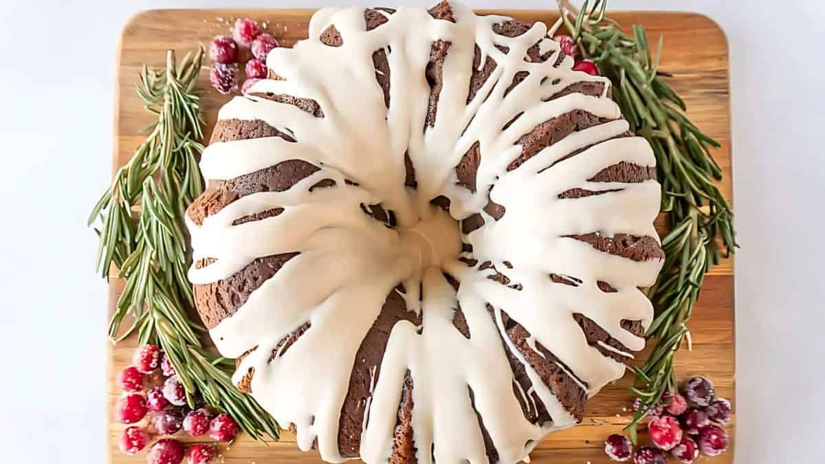 A Bundt cake with white icing drizzled on top sits on a wooden board. It is garnished with rosemary sprigs and sugared cranberries, creating a festive presentation.