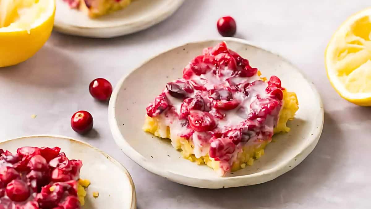 A dessert square with a creamy topping mixed with cranberries is served on a small plate. There are whole cranberries and a lemon half in the background on a light-colored surface.