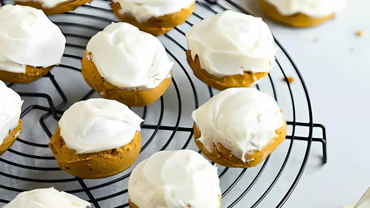 Pumpkin cookies topped with white frosting are cooling on a round wire rack. The cookies have a soft, round shape, and the frosting appears creamy and smooth. The background is a light surface.