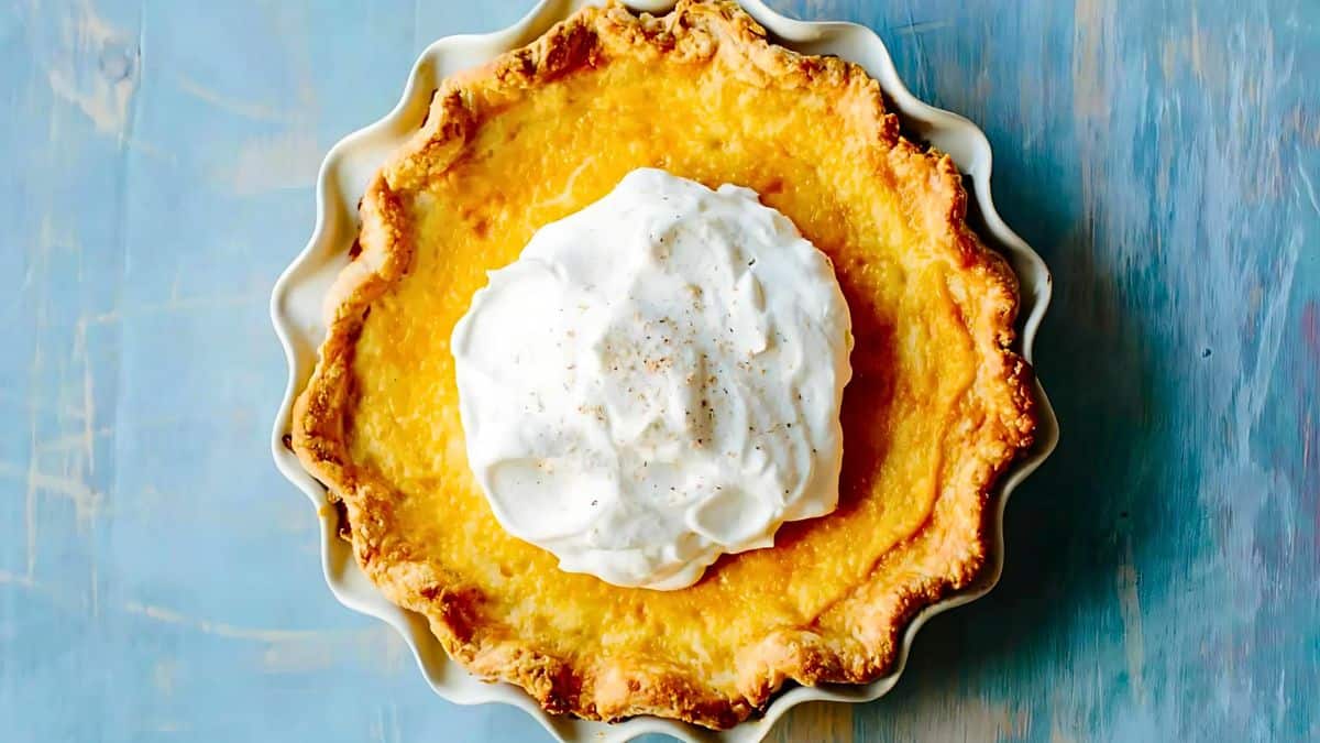 A golden brown pie with a flaky crust is topped with a generous dollop of whipped cream, set on a light blue background.